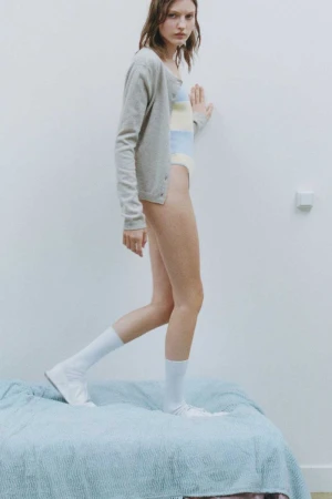 Model wearing a grey cardigan and white socks standing on a blue bed against a white wall for Claudie Pierlot.