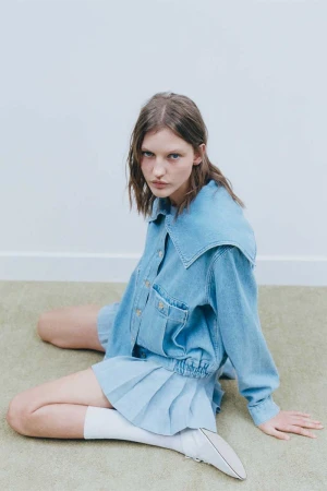 Model Nadine Kirilova wearing denim jacket and pleated skirt seated on floor for Claudie Pierlot campaign