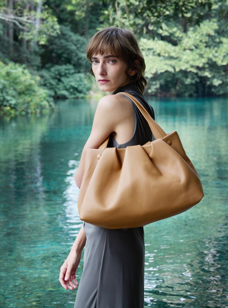 Model in grey dress carrying a large tan leather shoulder bag outdoors near a tranquil river set against a lush green forest background.