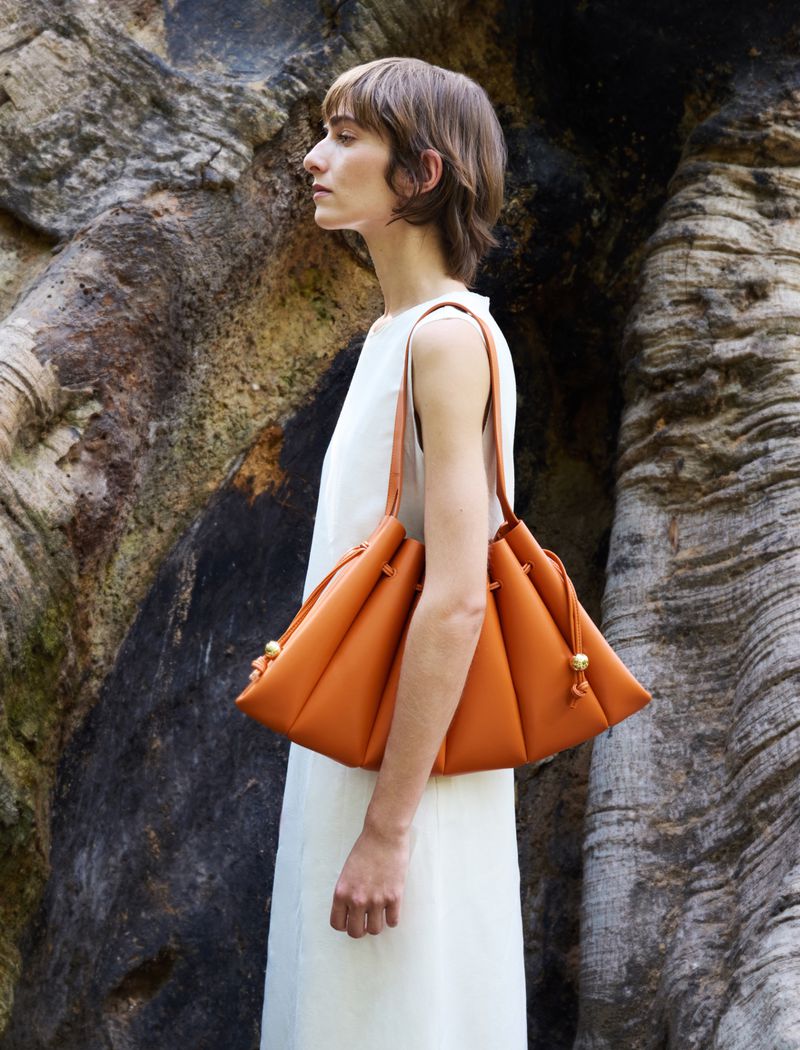 Model in white dress holding a pleated tan leather shoulder bag against a gnarled tree trunk