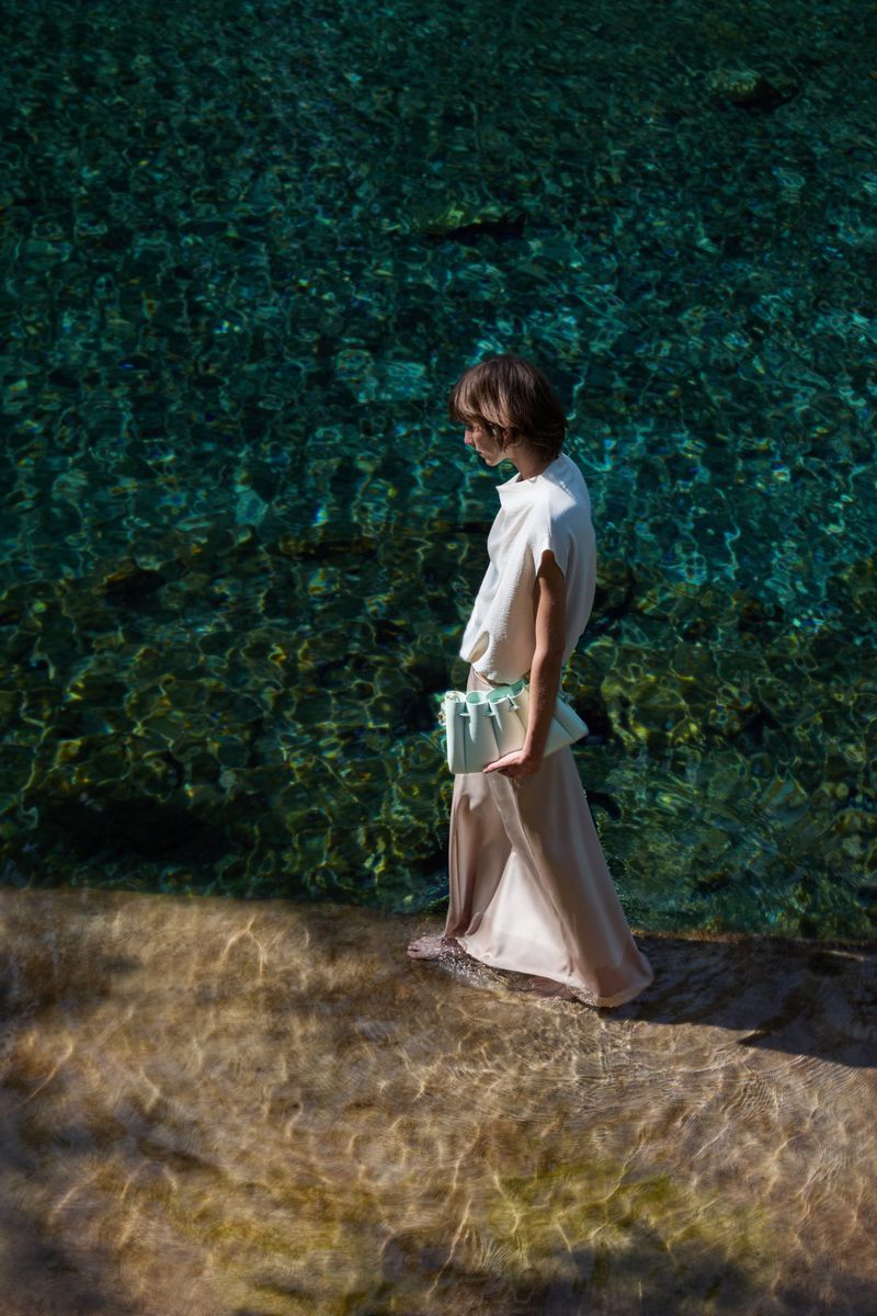 Woman in a flowing dress and a clutch stands near clear, shallow water with sunlight reflecting off the ripples and the sandy bottom.