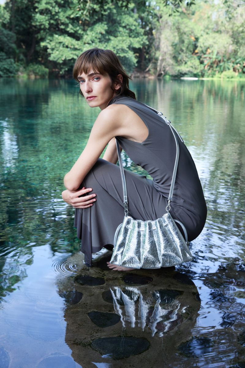 Woman in gray dress with silver purse crouching near lake surrounded by trees, creating a serene and fashionable outdoor scene.