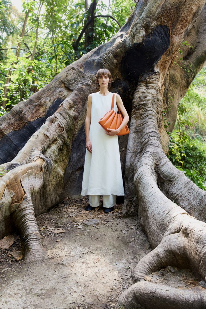 Woman poses by a tree in a sleeveless white dress, carrying a tan purse in a natural, sunlit outdoor setting with lush green foliage.