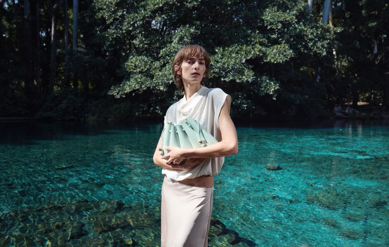 Model standing waist-deep by a crystal-clear blue spring, holding a pale pleated handbag against a lush forest backdrop.
