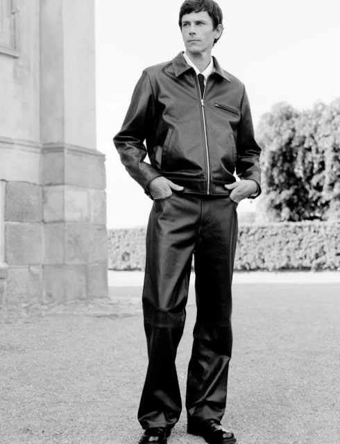 Man poses in black leather jacket and pants outside a building with trimmed bushes in the background, shot in black and white.