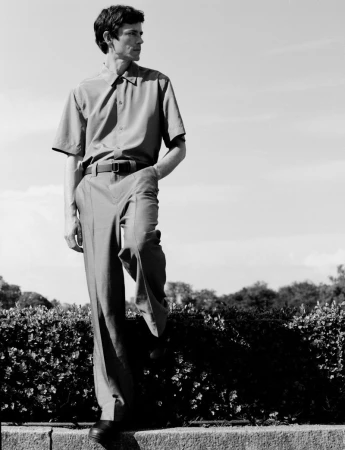 Monochrome full-length portrait of a male model standing outdoors wearing a short-sleeve button-down shirt and trousers.