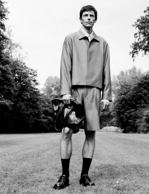 Black and white shot of a male model posing in a matching jacket and shorts outfit, holding a bag, on a grassy lawn.