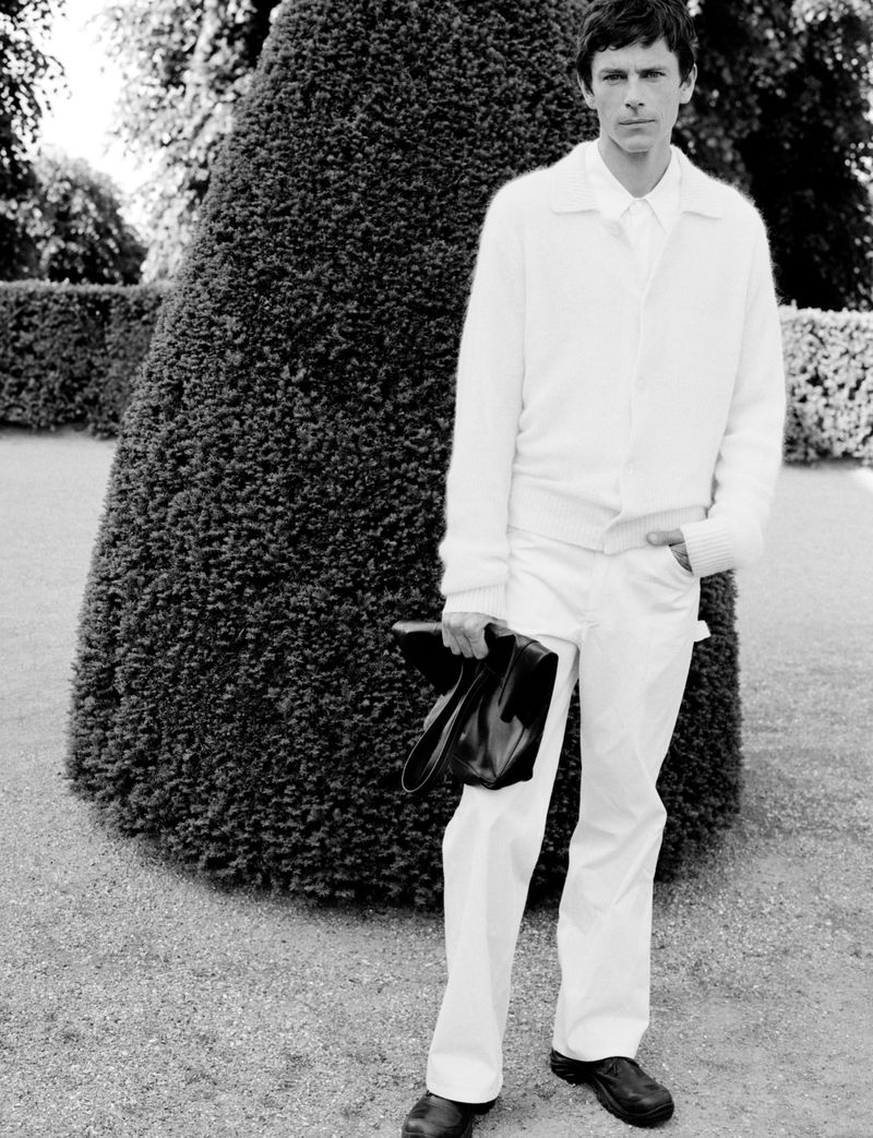 Model Jeremy Young wearing a white cardigan and trousers standing outdoors in a garden for Berner Kühl Spring Summer 2025 Campaign