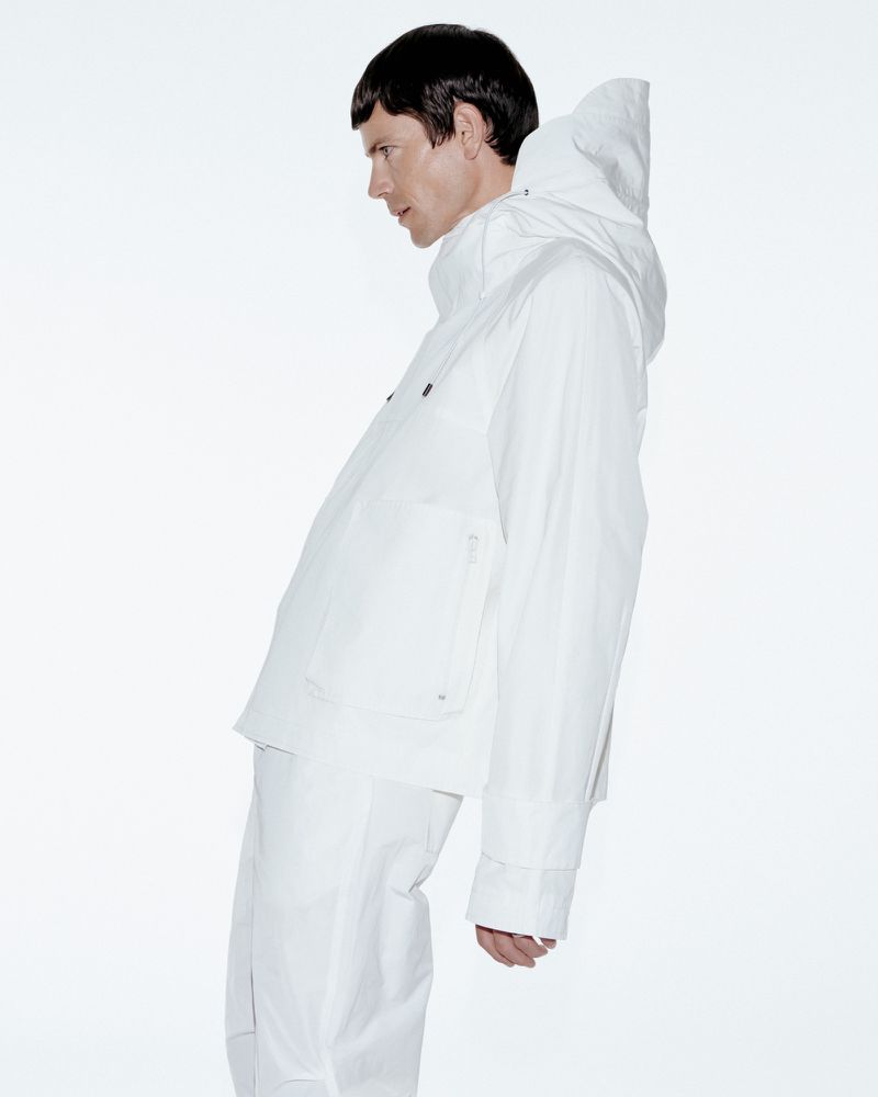 Profile view of a person in a white hooded jacket and pants against a bright white background in a fashion or editorial setting.