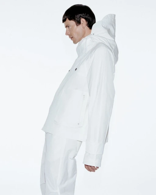 Profile view of a person in a white hooded jacket and pants against a bright white background in a fashion or editorial setting.