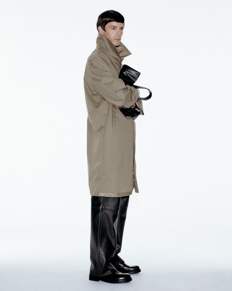A slender adult male stands in a studio wearing a tan trench coat, black leather boots, and gloves while holding a dark bag.