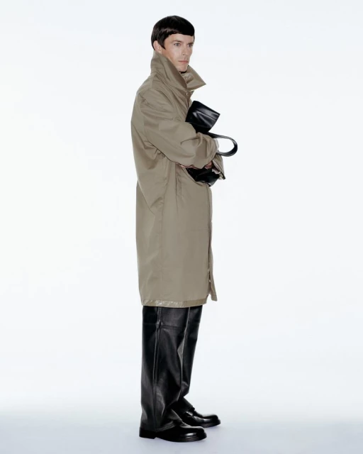A slender adult male stands in a studio wearing a tan trench coat, black leather boots, and gloves while holding a dark bag.