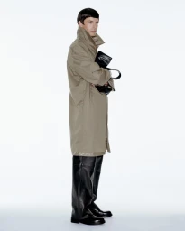 Model wearing a long beige trench coat and black trousers holding a black handbag against a white studio background.