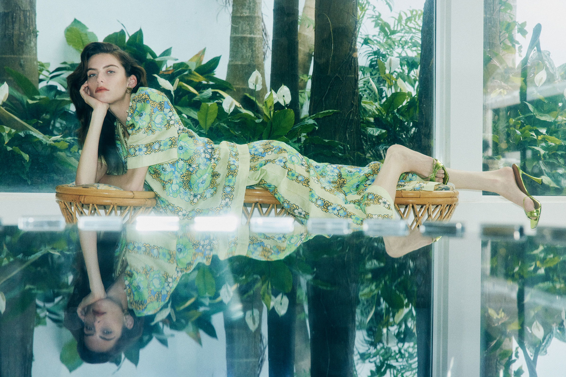 Woman in patterned yellow loungewear reclines on a woven stool on a reflective surface, surrounded by tropical greenery and natural light.