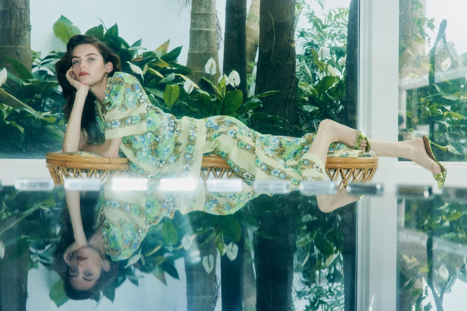 Woman in patterned yellow loungewear reclines on a woven stool on a reflective surface, surrounded by tropical greenery and natural light.