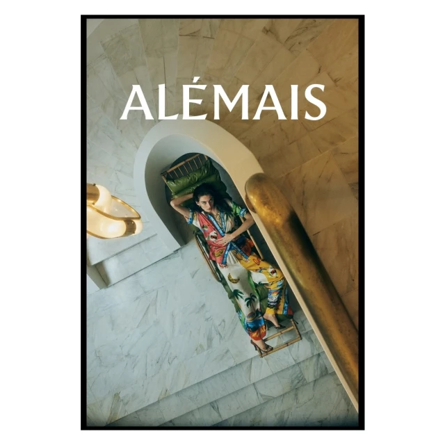 A woman lounges on a chaise lounge on a marble staircase next to a bronze railing. An ad features the brand name, Alémais.