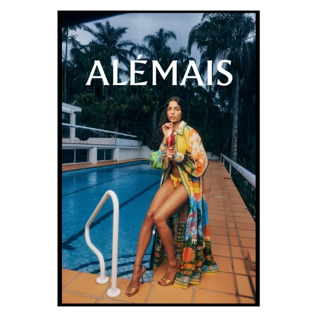 A model poses next to a pool in a multicolored robe with a drink; palm trees and architecture in the background; fashion shoot.