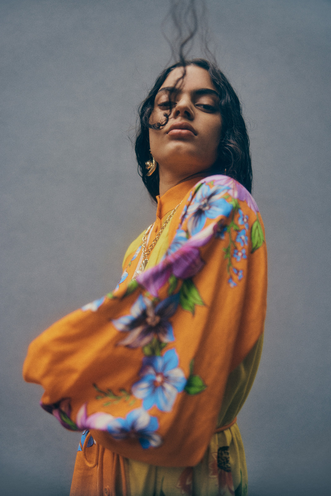 Portrait of a fashionable young adult woman wearing a floral patterned orange dress, gold jewelry, looking at the camera with neutral expression.