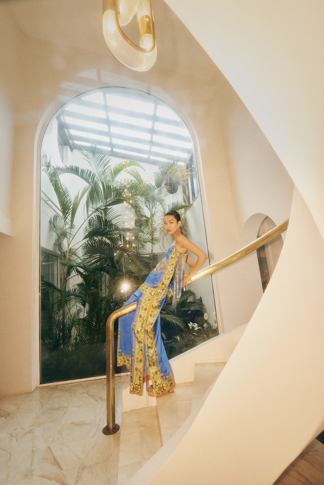 Woman in blue-and-yellow dress on stairs with brass railing, indoor plants and natural light, modern architectural details.