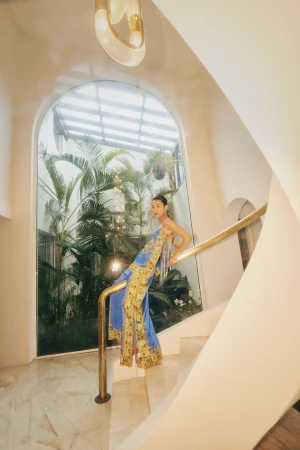 Woman in blue-and-yellow dress on stairs with brass railing, indoor plants and natural light, modern architectural details.