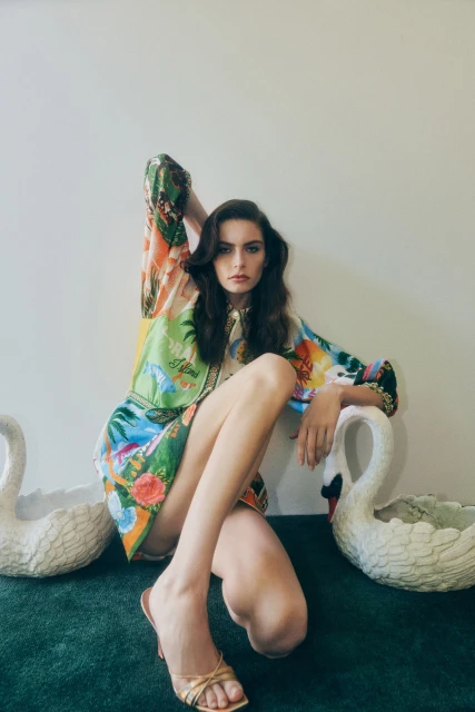 A woman poses in a patterned dress with swan planters indoors, showcasing fashion photography with natural light against a white wall.