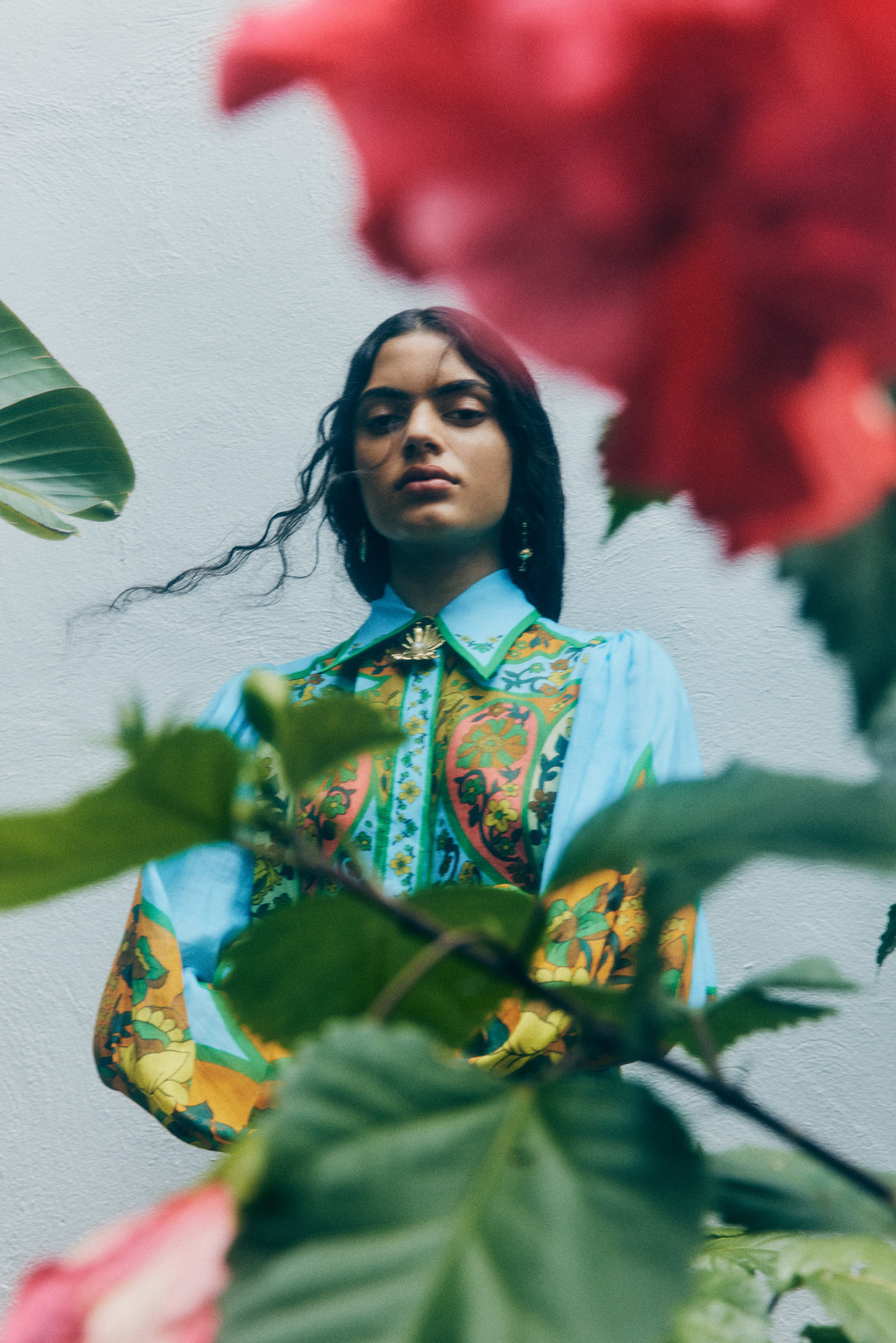 Woman in a patterned shirt stands against a white wall, framed by vibrant red and green plants, illuminated by natural light.