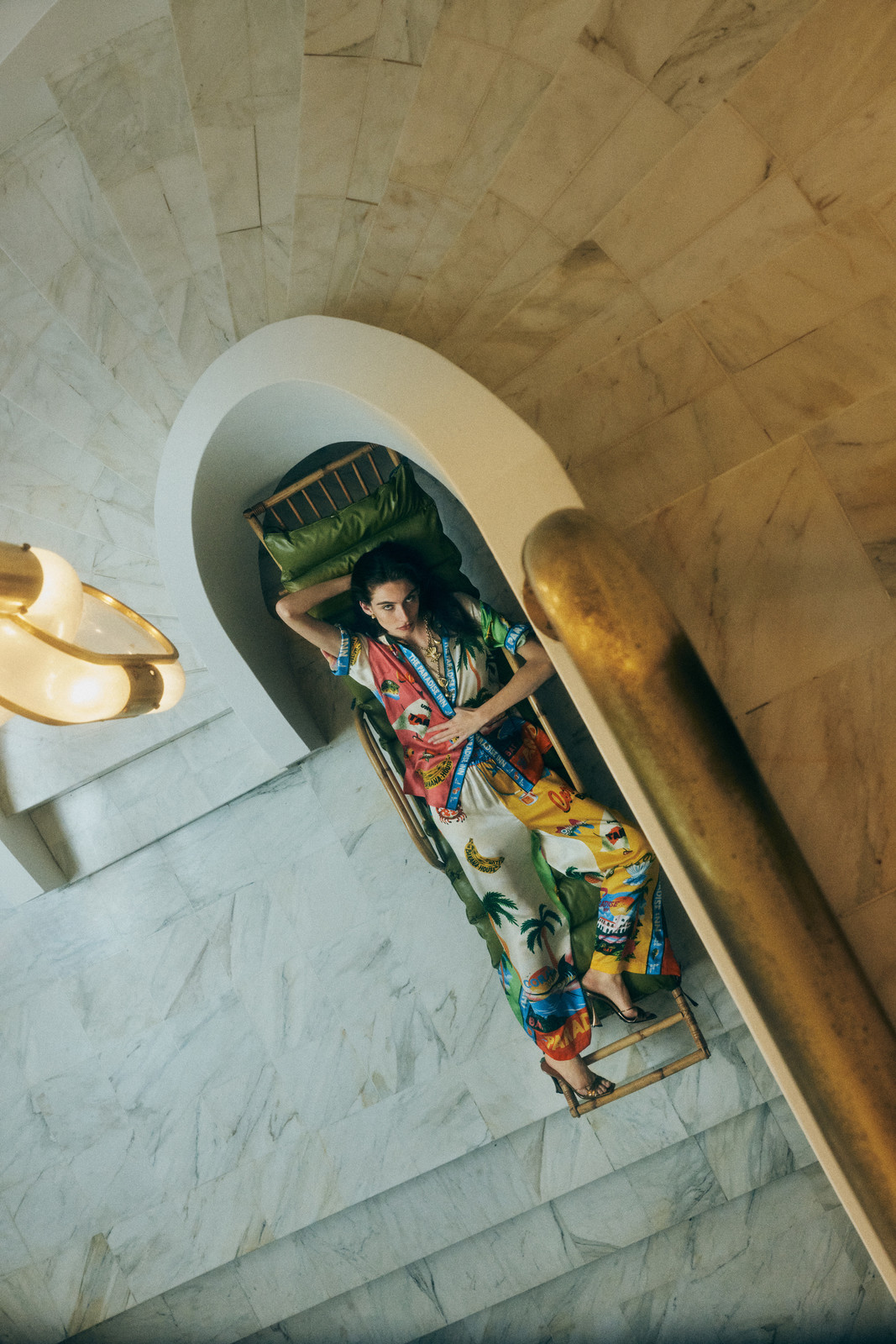 Woman in patterned pajamas lies on a bamboo lounge on marble stairs indoors with a curved entryway and brass chandelier, overhead angle.