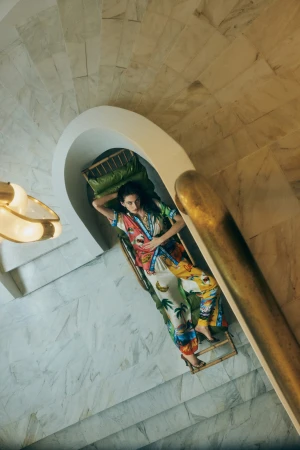 Woman in patterned pajamas lies on a bamboo lounge on marble stairs indoors with a curved entryway and brass chandelier, overhead angle.