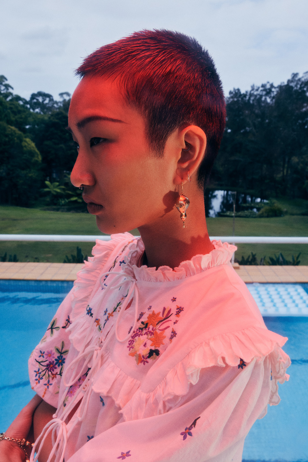 Profile view of a person with short hair wearing a floral top outdoors by a pool. The image features soft, diffused lighting.