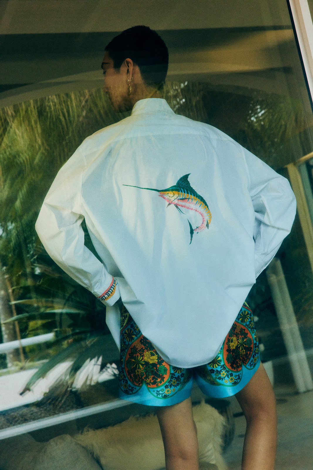 Back view of a stylish adult wearing a white button-down shirt with a marlin print and patterned turquoise shorts outdoors.