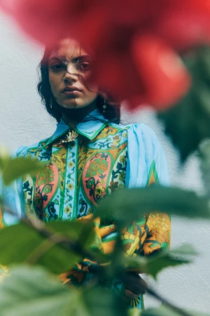 Model in a patterned blue and yellow blouse viewed through out-of-focus red flowers and green leaves in the foreground.