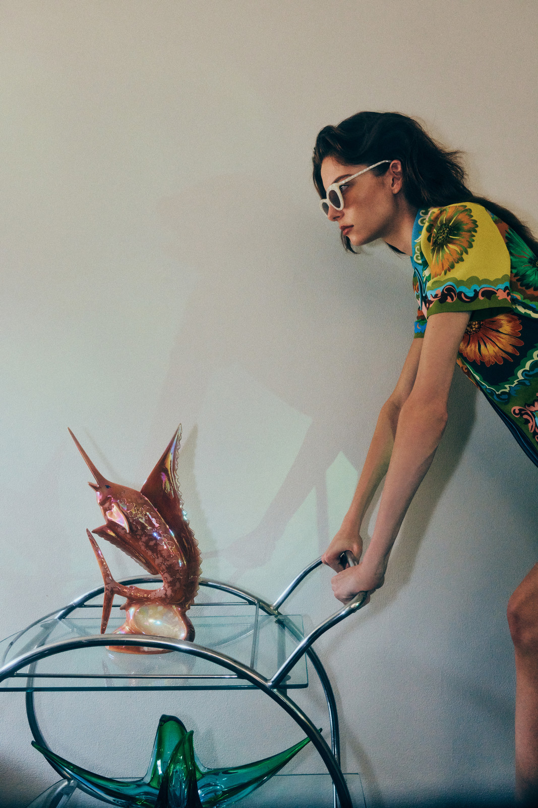 Woman wearing sunglasses and printed dress pushes a bar cart holding a glass fish sculpture against a neutral background indoors.