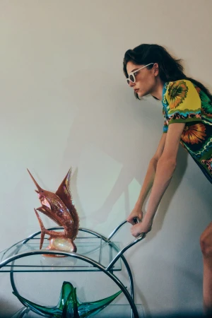 Woman wearing sunglasses and printed dress pushes a bar cart holding a glass fish sculpture against a neutral background indoors.