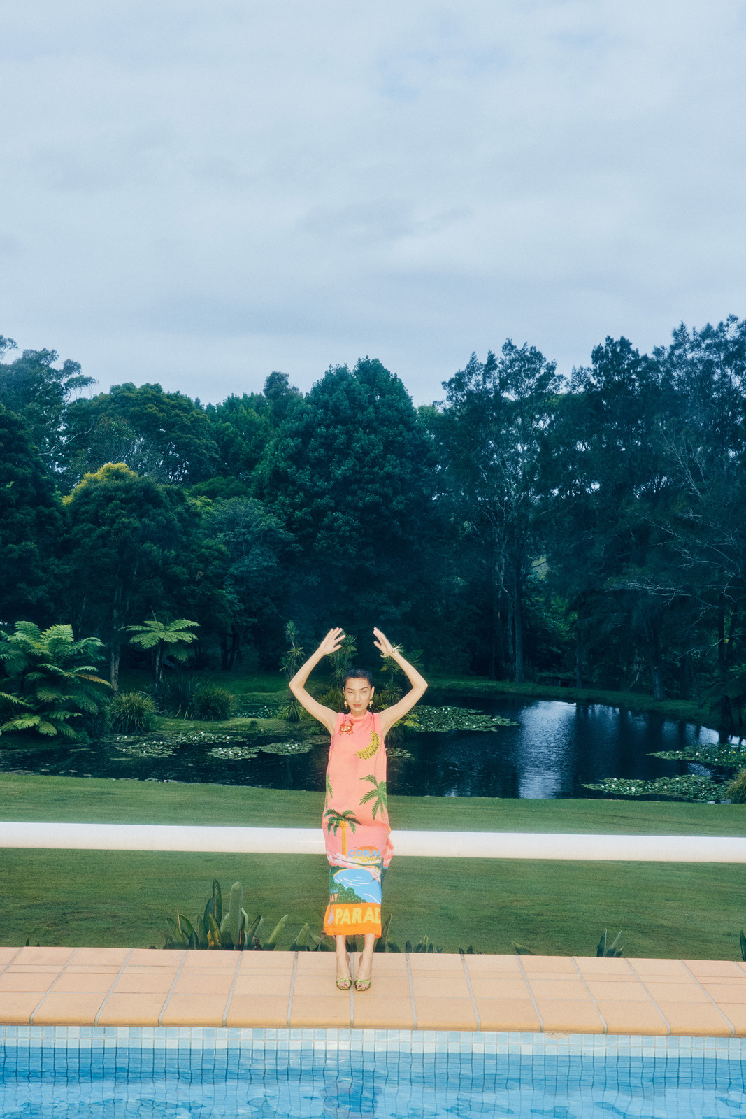Woman in a tropical print dress stands near a pool with hands raised. Trees and a pond are in the background under a cloudy sky.