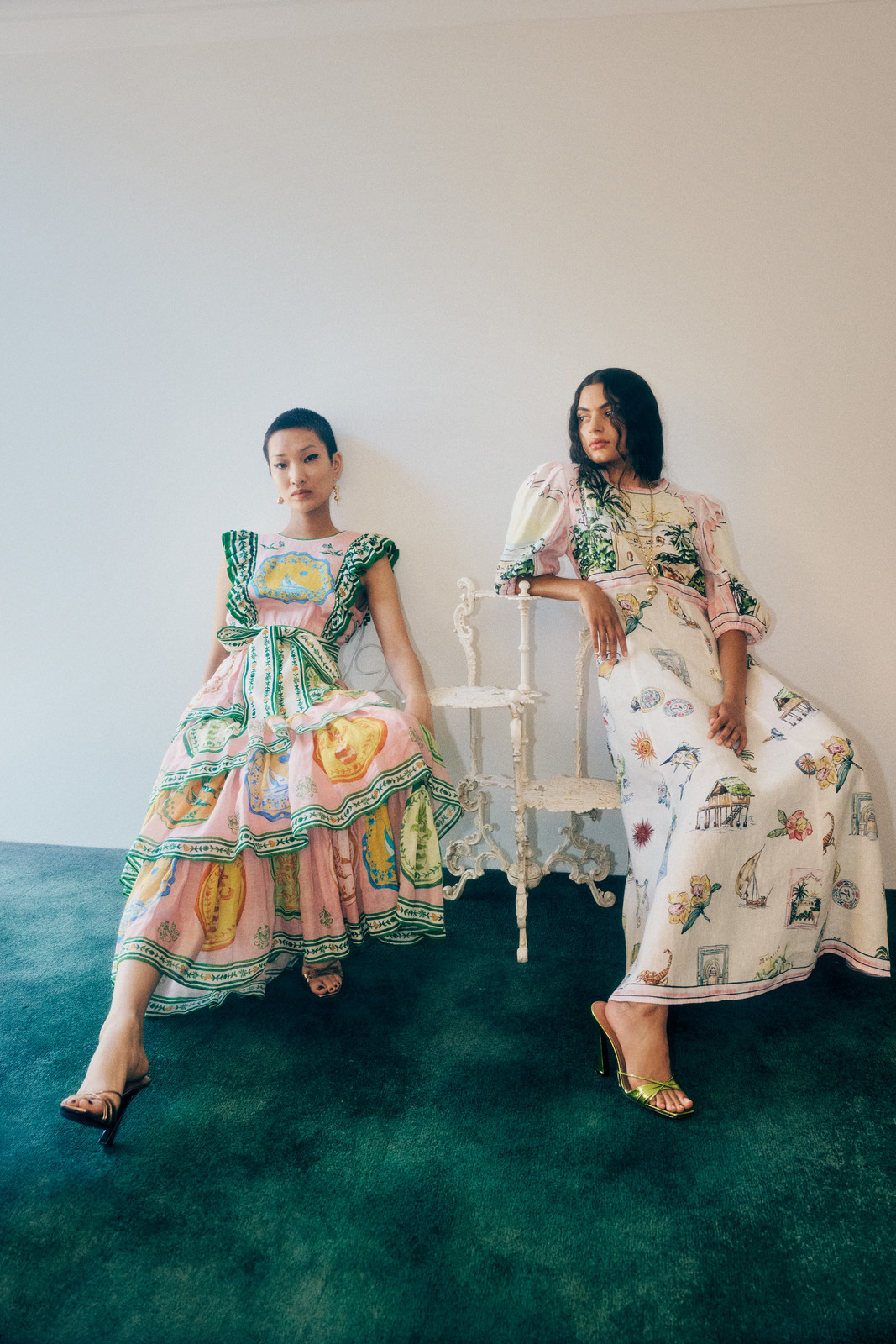 Two women pose indoors wearing patterned dresses and heels, seated against a white wall on a green textured floor. Fashion Editorial.