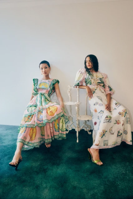 Two women pose indoors wearing patterned dresses and heels, seated against a white wall on a green textured floor. Fashion Editorial.