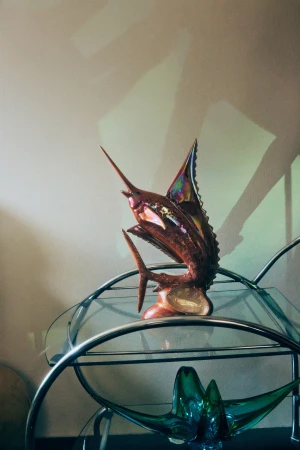 Bronze sailfish sculpture on a glass and metal stand, showcasing intricate craftsmanship and reflective surfaces against a neutral backdrop.