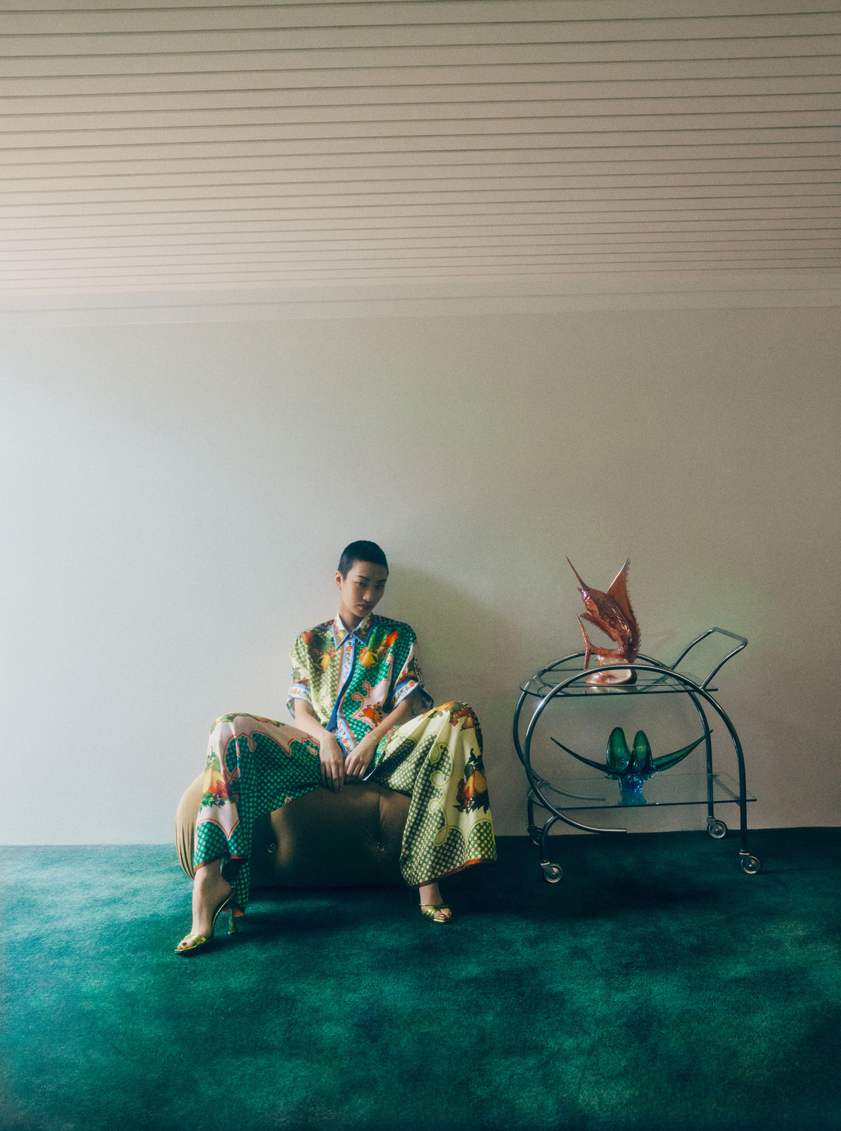 Woman in patterned co-ord set seated next to metal bar cart with abstract sculpture in a sparse room with green carpet.