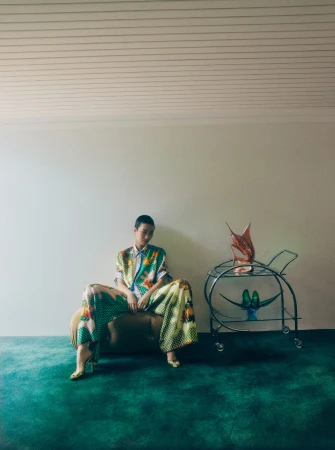 Woman in patterned co-ord set seated next to metal bar cart with abstract sculpture in a sparse room with green carpet.