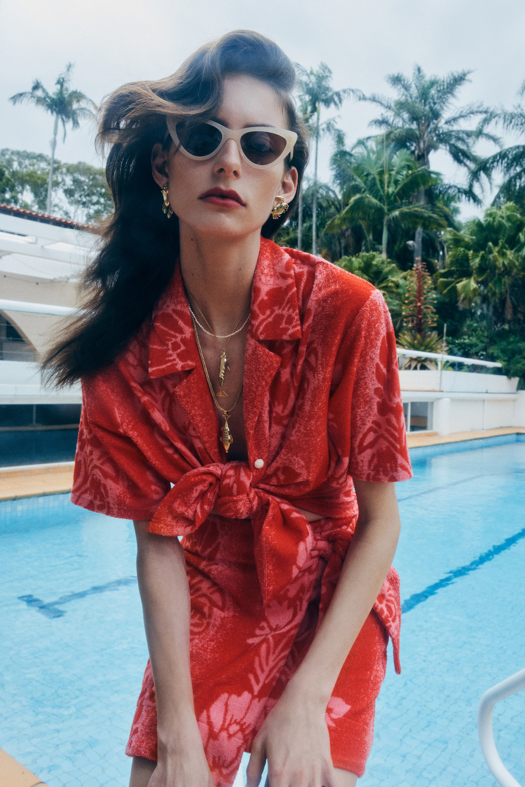 A woman poses in a red floral outfit with palm trees and a pool in the background, wearing sunglasses and jewelry for a fashion shot.