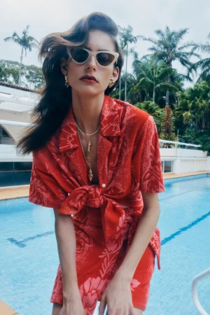 A woman poses in a red floral outfit with palm trees and a pool in the background, wearing sunglasses and jewelry for a fashion shot.