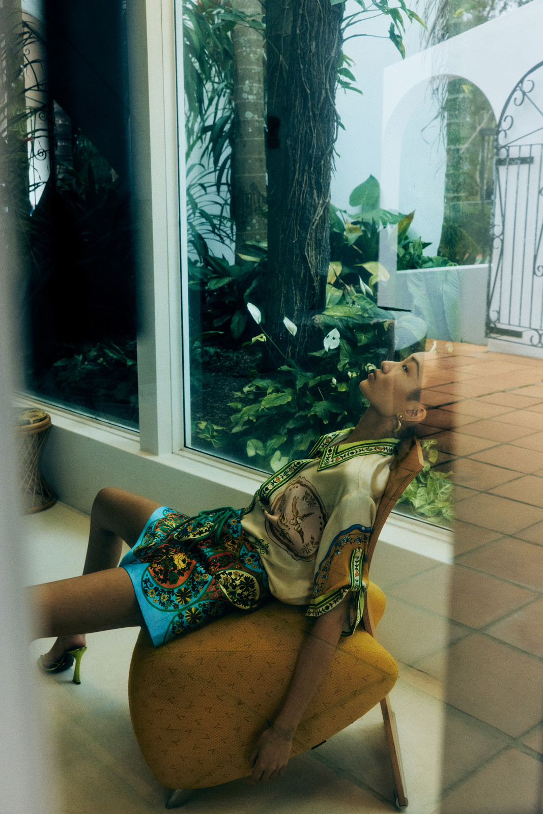 Adult female model rests on yellow chair indoors. She wears a printed top and shorts with green high heels by a large window.