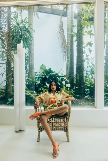 A woman in a tropical-patterned shirt sits in a wicker chair in front of large windows and greenery, wearing orange gladiator sandals.