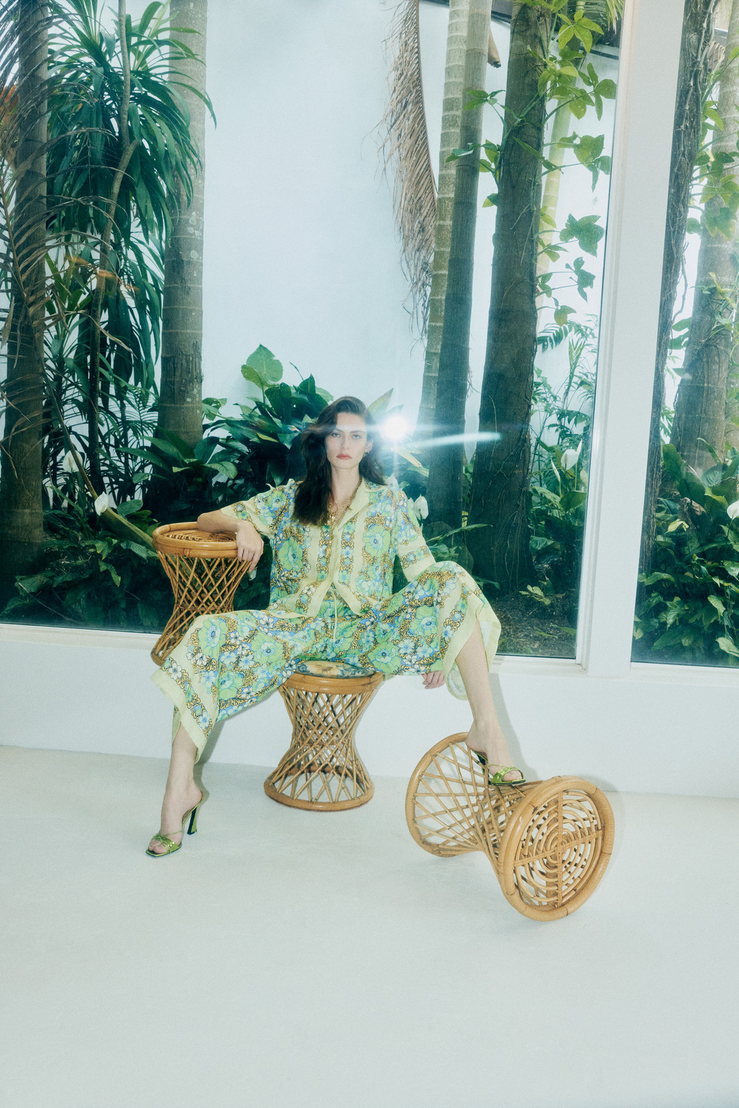A woman wearing a yellow and green patterned pajama set sitting on a wicker stool with tropical plants and light filtering through the window.