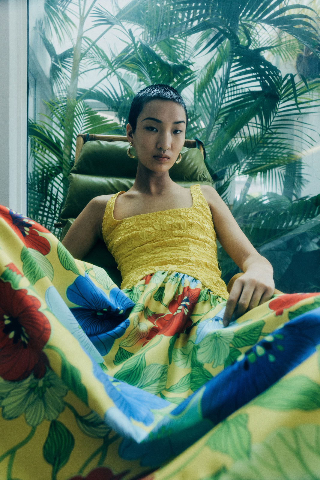 Model with short dark hair seated in a wicker chair wearing a yellow patterned floral dress against a tropical palm background.