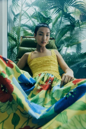 Model with short dark hair seated in a wicker chair wearing a yellow patterned floral dress against a tropical palm background.