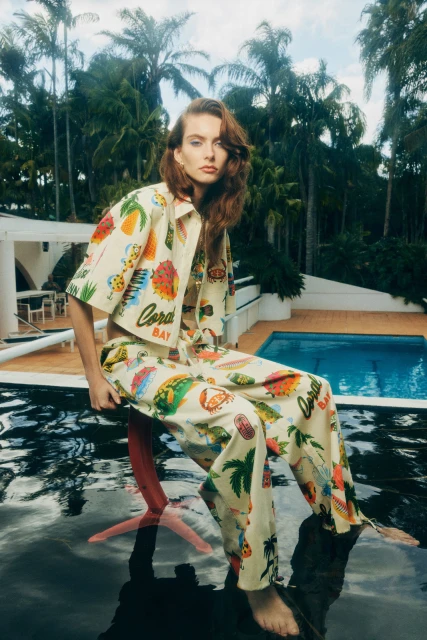 A redhead woman in a patterned two-piece outfit sits on a chair, reflecting in a wet surface, with a pool and palm trees.