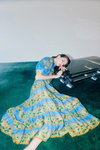 Fashion shot of a woman reclining on a green surface wearing a blue and green floral patterned dress in an indoor setting.