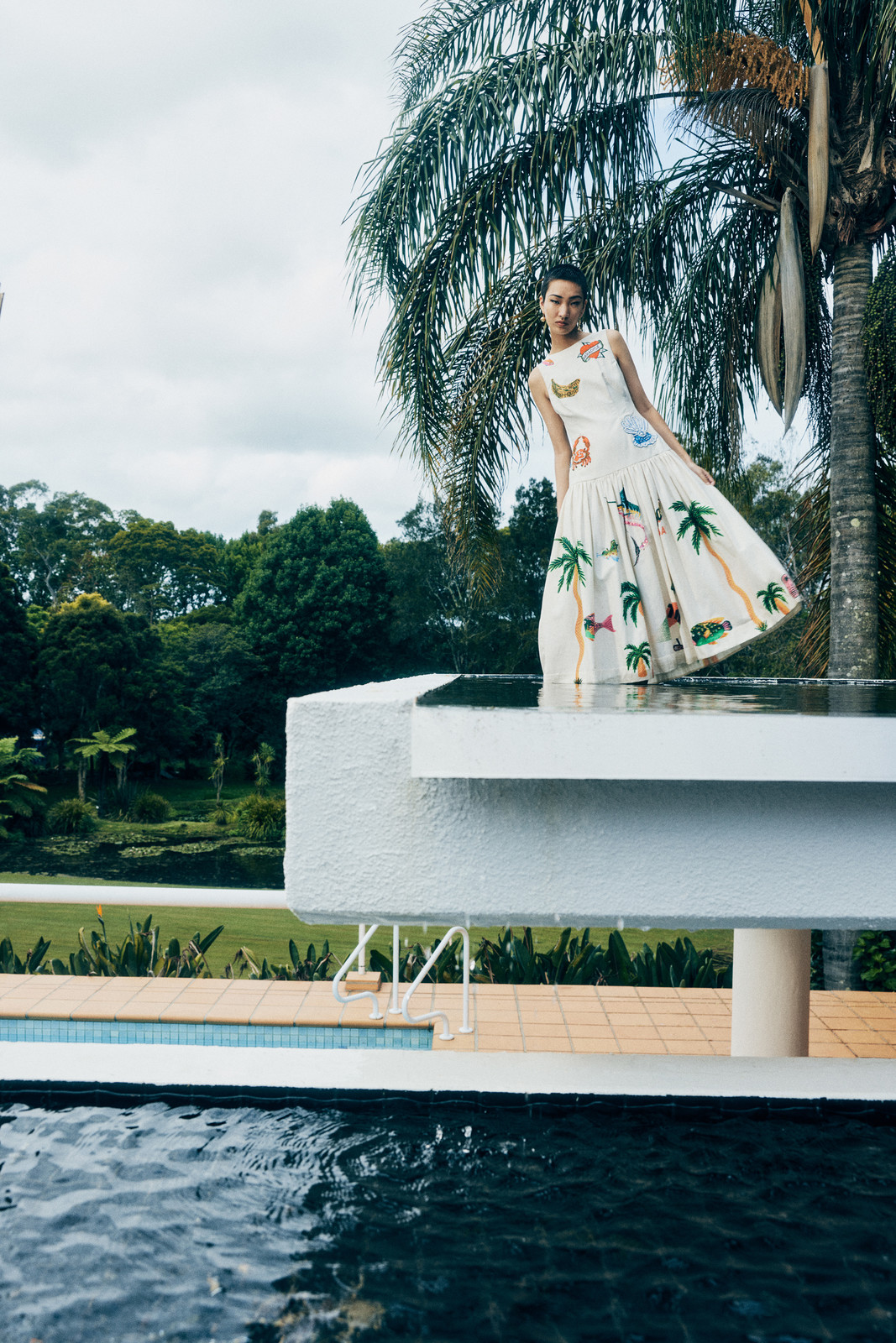 Woman in a printed white sundress posing on a poolside ledge with palm trees and greenery in the background under a cloudy sky.