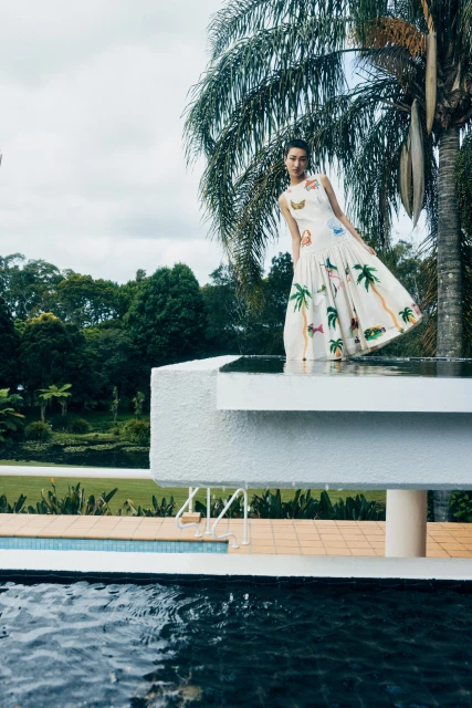 Woman in a printed white sundress posing on a poolside ledge with palm trees and greenery in the background under a cloudy sky.
